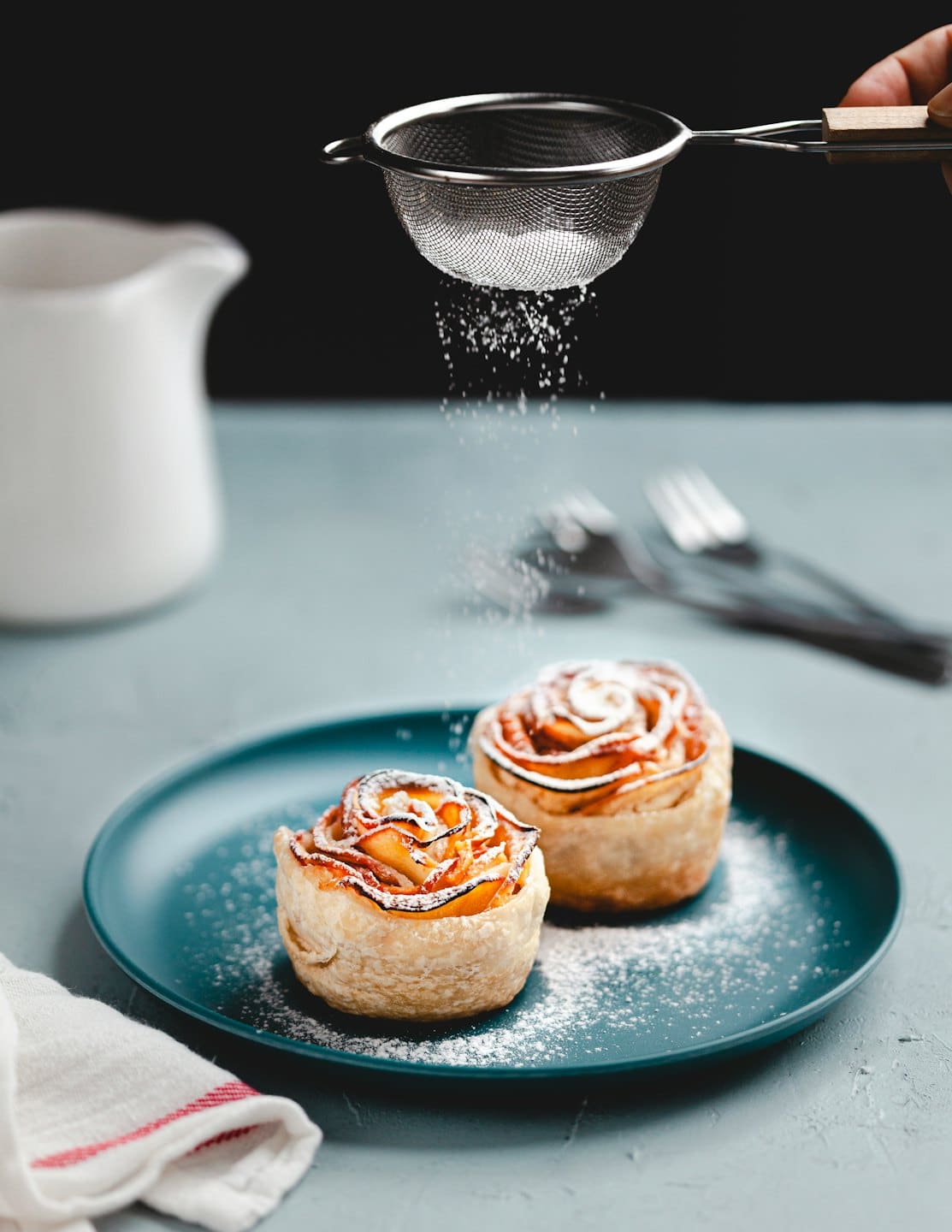 Manos espolvoreando azúcar glas sobre tartas de manzana en forma de rosa sobre un plato turquesa.